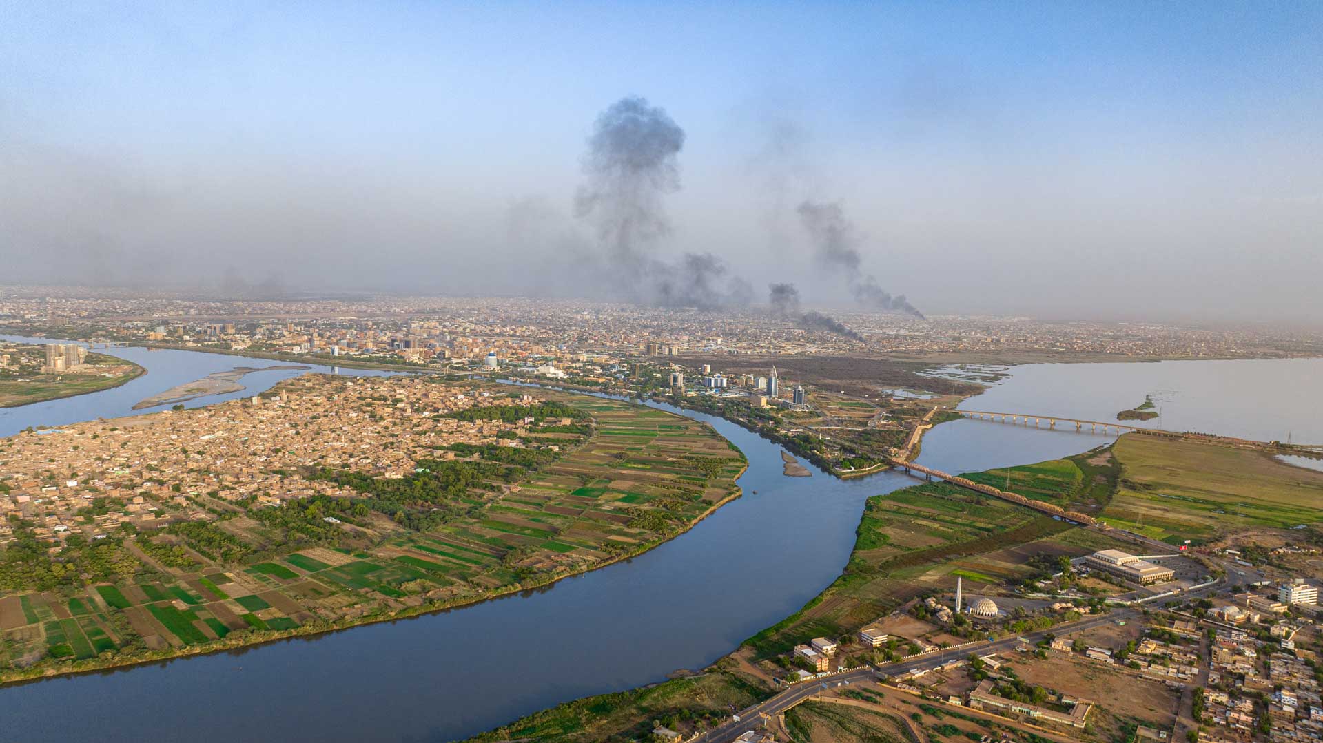 Drone footage looking down over and south towards the Khartoum area. The Y shaped confluence of the Blue and White Niles cuts the centre of the picture, to the left and right the banks have narrow green strips of field, backing onto housing. In the distance the centre of Khartoum is built up, black smoke rises from two large fires, the hazy sky gives way to blue towards the top of the image.