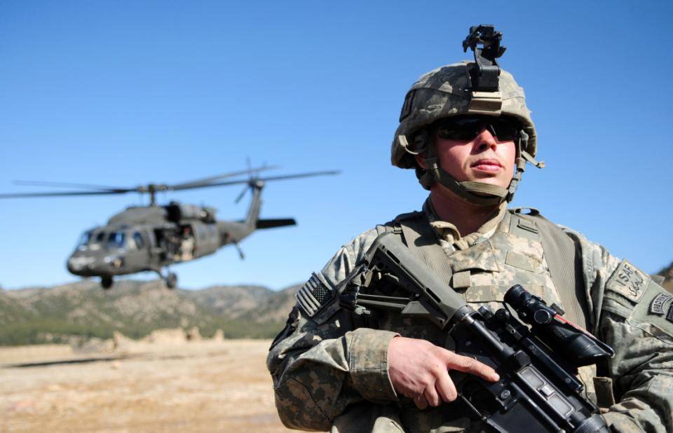 A US soldier in Afghanistan stands with agun, while a Blackhawk helicopter lands behind him.