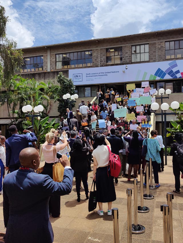 Civil society representatives hold up placards on steps outside UNEA-6 in a silent protect on the environmental consequences of conflicts.