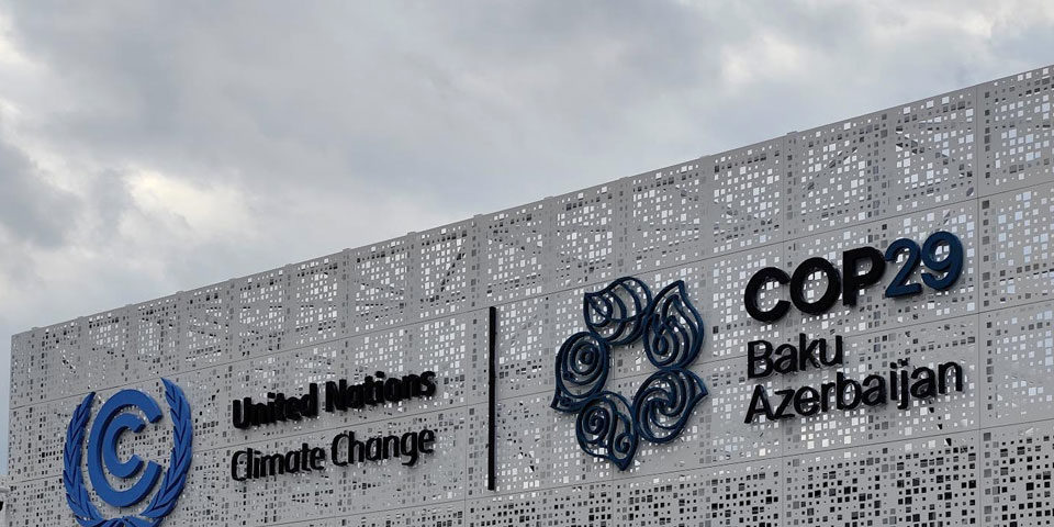 COP29_header_960 The entrance to the building that will be home to COP29 showing the UNFCCC logo and COP29's logo against a grey sky.