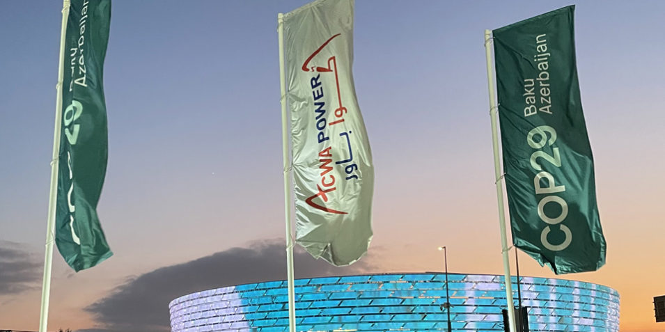 cop29_centre_960 In the foreground, three banners promoting COP29, behind them a large modern stadium is lit up in blue and white lights as the sun sets behind it.