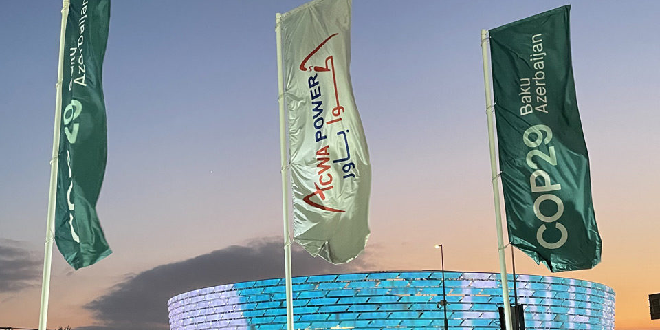 cop29_centre_960 In the foreground, three banners promoting COP29, behind them a large modern stadium is lit up in blue and white lights as the sun sets behind it.