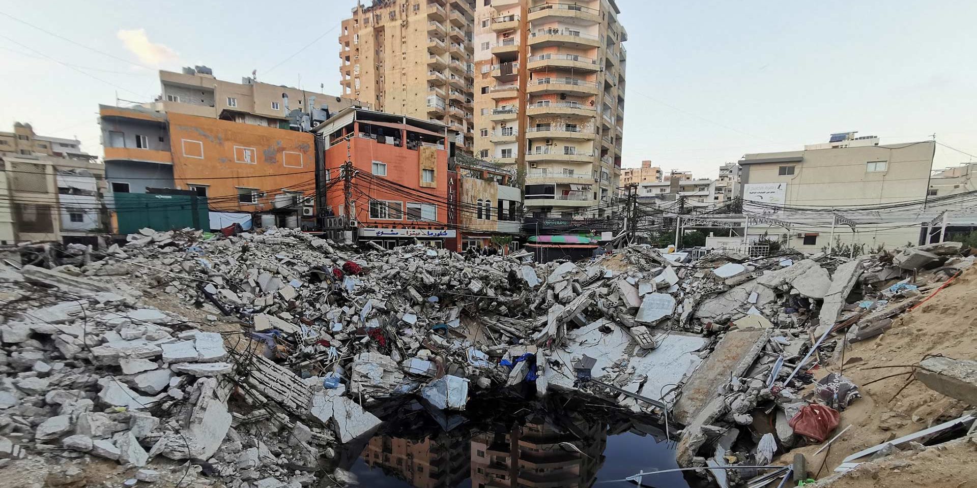 crater_clo_1920 A deep crater in the foreground is half full of water, half of the crater sides are covered in the debris from a demolished building, behind this a row or three story buildings are painted orange and pink, behind them two 10 storey apartment blocks.