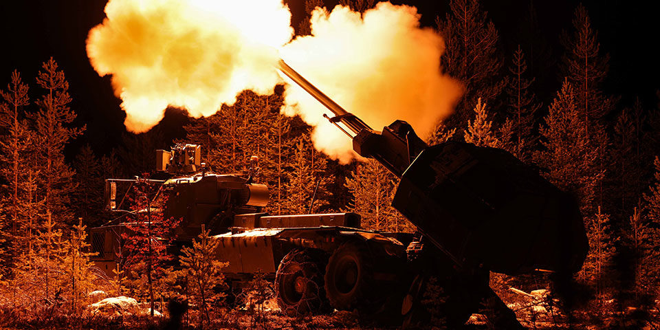 Live-firing-UKMOD_finland_960 This night firing image shows the dark silhouette of the self-propelled artillery in a wooded area. The trees and part so the vehicle are lit up from the flash of the gun muzzle, clouds of gas are coming from each side of the gun and are illuminated white and orange.