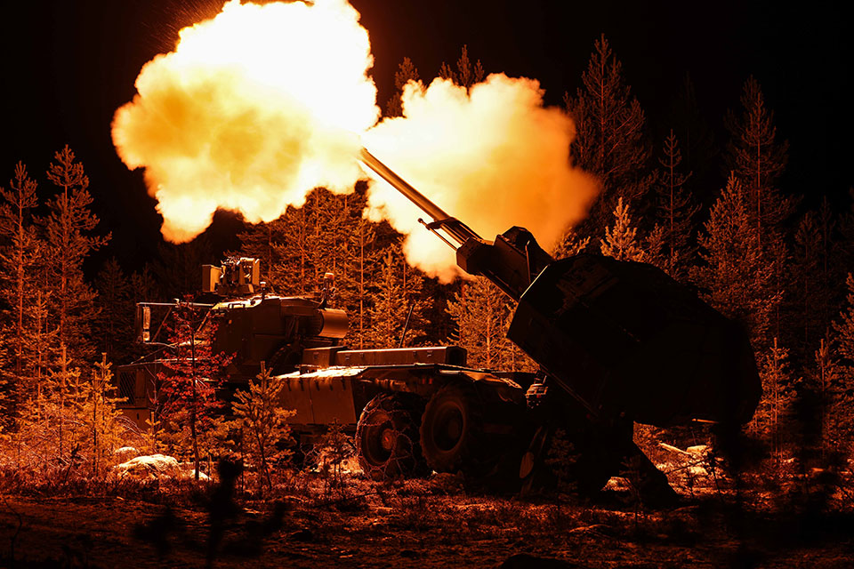 This night firing image shows the dark silhouette of the self-propelled artillery in a wooded area. The trees and part so the vehicle are lit up from the flash of the gun muzzle, clouds of gas are coming from each side of the gun and are illuminated white and orange.