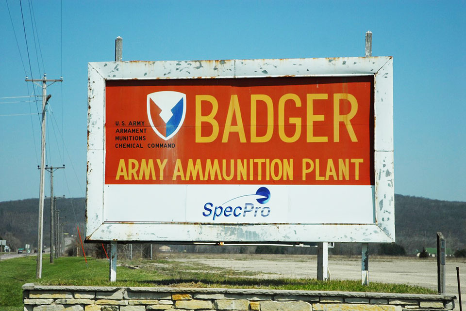 A large sign against a blue sky, the sign reads "Badger Army Ammunition Plant" in tal yellow lettering on a red background.