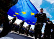 A photo from ground level looks up at soldiers carrying the EU flag in a parade, the flag is lit up by the blue sky and sunny weather above.