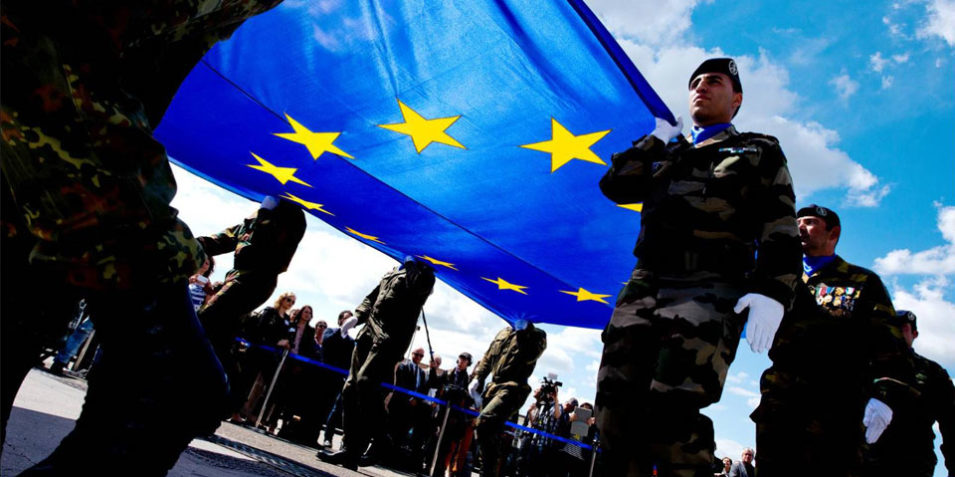EU_flag_960 A photo from ground level looks up at soldiers carrying the EU flag in a parade, the flag is lit up by the blue sky and sunny weather above.