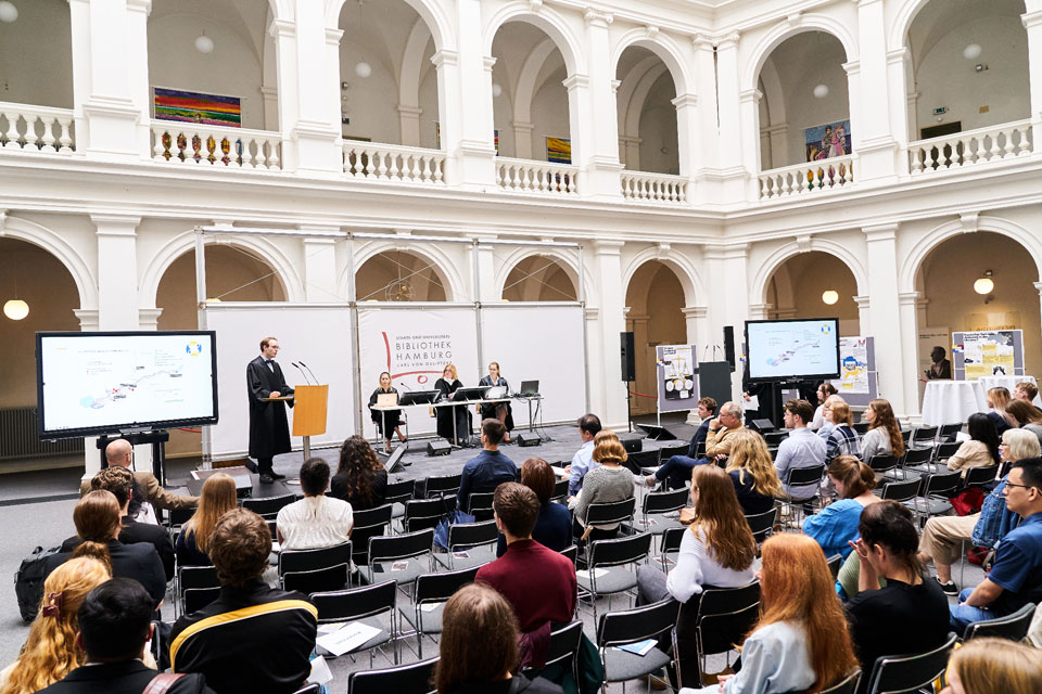 The grand central atrium of the Bibliotek Hamburg. A seated audience watches a low stage, on it a man in legal robes stands at a podium addressing the audience. To his right on teh stage, three black robed judges sit listening. Behind, two floors of colonnades in white marble rise above the scene.