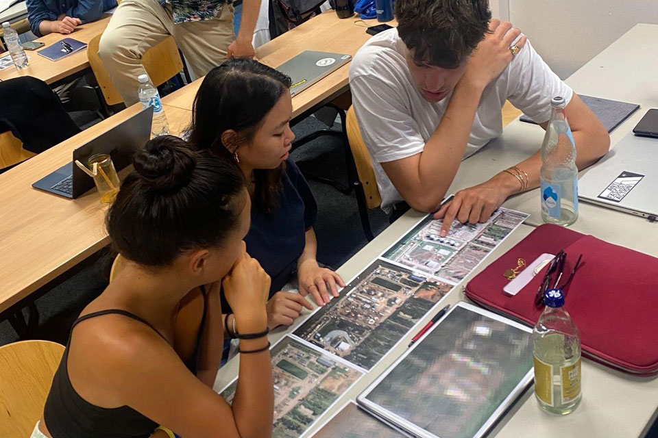 Three students sit at a desk looking at blown up satellite images of facilities in Ukraine, they are deep in conversation.