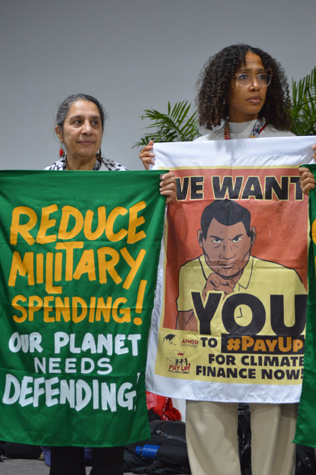 Two female campaigners stand holding banners at COP29, the green banner on the left says REDUCE MILITARY SPENDING OUR PLANET NEEDS DEFENDING in yellow and white letters