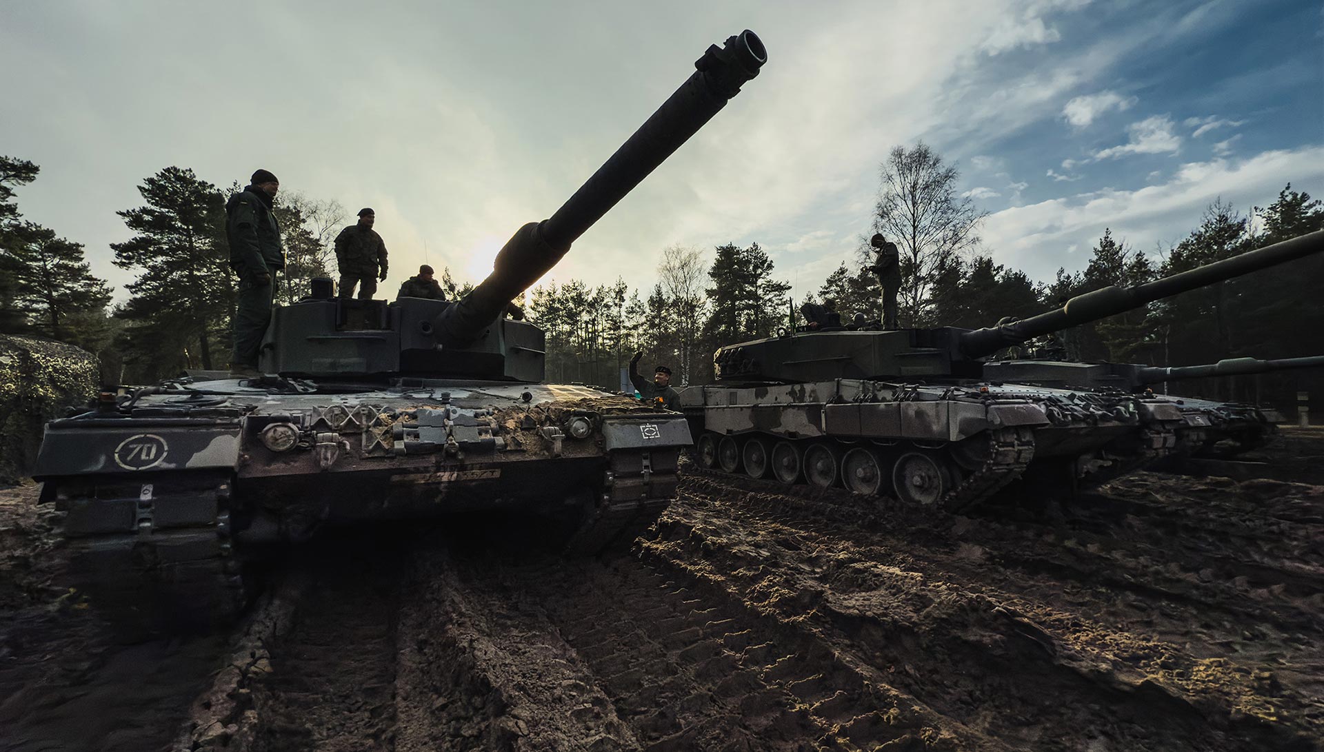 Three parked leopard battle tanks, soldiers stand on the two nearest, the ground is muddy and churned by tank tracks, behind is woodland and a clear sky.