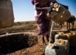 In teh left of the image a circular opening to a water well can be seen, the land beyond is arid brown and green under a blue sky. The lower two thirds of a woman can be seen in the right of the image, she is pouring water into jerry cans.