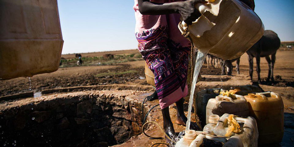 sudan_unep_project_960 In teh left of the image a circular opening to a water well can be seen, the land beyond is arid brown and green under a blue sky. The lower two thirds of a woman can be seen in the right of the image, she is pouring water into jerry cans.