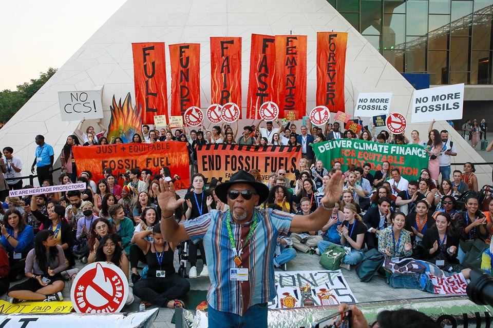 A photo looking down on a group of around 100 protestors at COP28, many different banners and slogans can be seen among the diverse crowd.