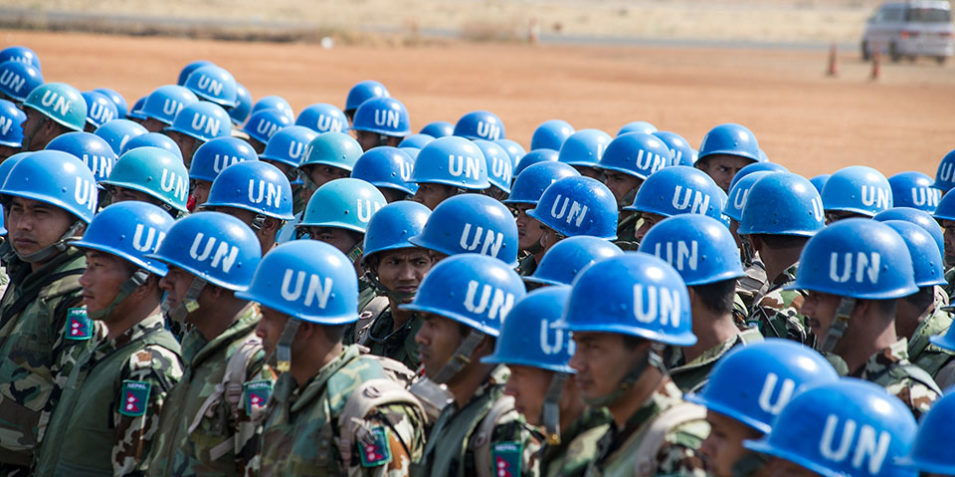 UN_blue_helmets_960 A crowd of UN Peacekeepers is visible in the foreground, wearing camouflage uniform and their distinctive blue helmets emblazoned with a white "UN". In the background brown arid desert. Around 40 troops are visible.
