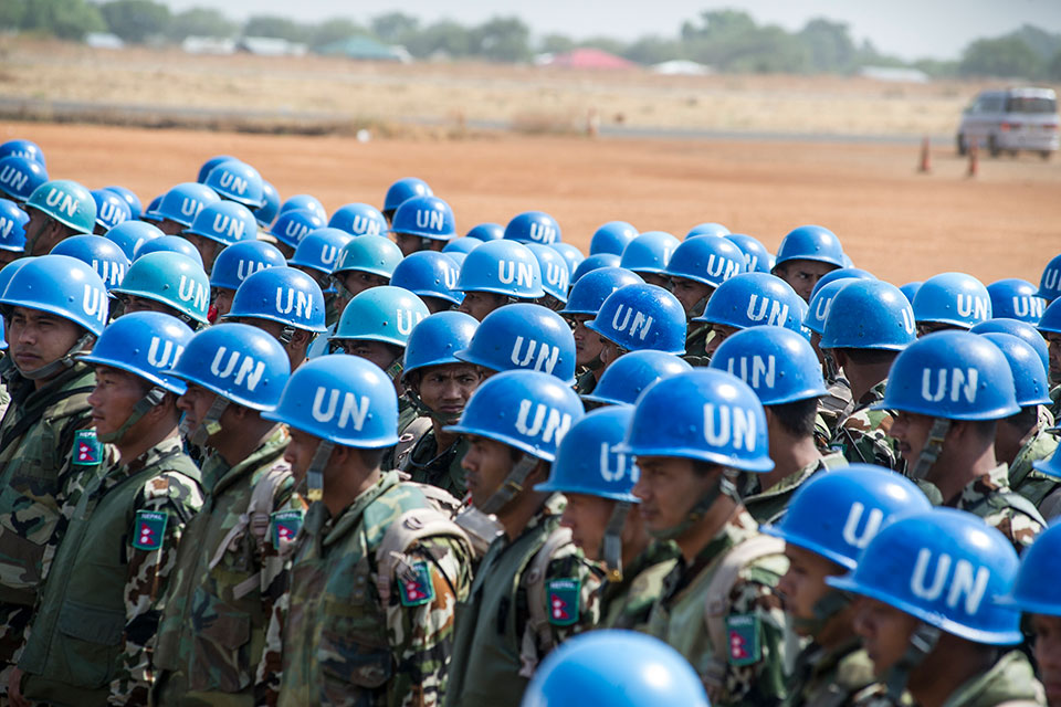 A crowd of UN Peacekeepers is visible in the foreground, wearing camouflage uniform and their distinctive blue helmets emblazoned with a white "UN". In the background brown arid desert. Around 40 troops are visible.