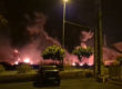 Grainy photo taken from a car park at night, in the foreground a parked car and grassy border with small trees, behind large fires are visible that uplight the bases of dark smoke clouds. A man looks on in the right hand corner.