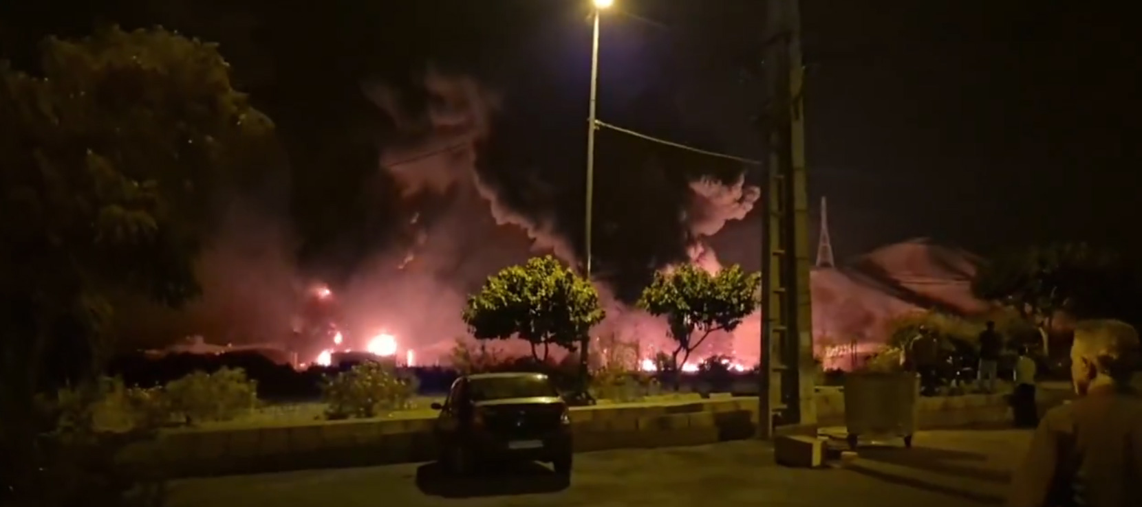 Grainy photo taken from a car park at night, in the foreground a parked car and grassy border with small trees, behind large fires are visible that uplight the bases of dark smoke clouds. A man looks on in the right hand corner.