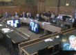 A wood-paneled parliamentary committee room, in which 10 people are seated in discussion around a large U-shaped table. Behind them to one side are five more people at two smaller tables. In the centre of the U, two large screens display three remote participants in the meeting. One of the women participants is also shown speaking in an inset in the corner of the image.