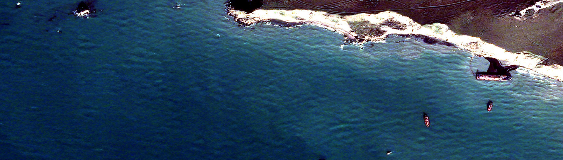 Satellite image of one of the wreck sites. Most of the image is the sea in shades or dark to mid blue, in the top right a coast line and white cliffs, in the far right of the image the lozenge like shape of a small tanker can be seen, just offshire, between it and the cliffs is a black slick of oil. Three smaller craft are offshore nearby.