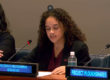 A young woman seated behind a blue digital sign reading the conflict and environment observatory. A conference microphone glows red.