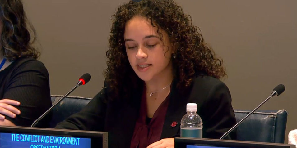 Malaika_960 A young woman seated behind a blue digital sign reading the conflict and environment observatory. A conference microphone glows red.