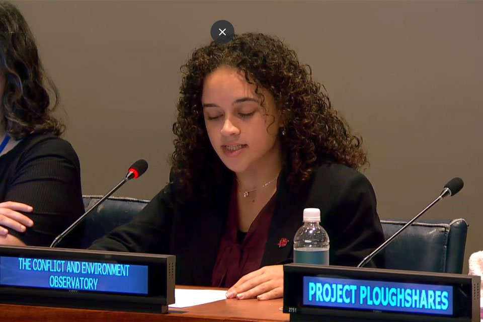 A young woman seated behind a blue digital sign reading the conflict and environment observatory. A conference microphone glows red.