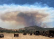 UK military personnel look on as a huge smoke plume rises from a distant ridgeline, a selection of army trucks and off road vehicles are in the foreground.