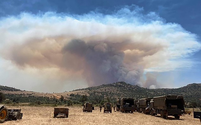 UK military personnel look on as a huge smoke plume rises from a distant ridgeline, a selection of army trucks and off road vehicles are in the foreground.