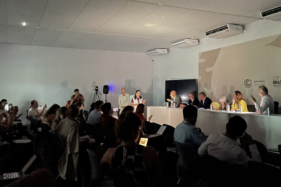 A partially lit room with shadows on the wall, to the right a six-person panel on a mounted stage are illuminated by phone torches as an audience to the left sites in partial darkness.