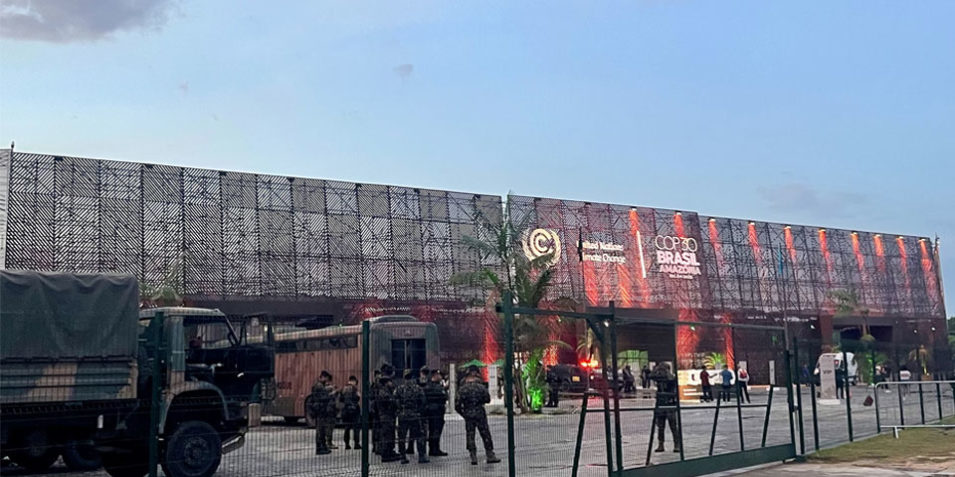 Behind a green mesh security fence military personnel can be seen in combat fatigures and berets. A troop carrying lorry is parked to the right. In the background the COP30 convention centre with its three storey mesh wall and UNFCCC logo.