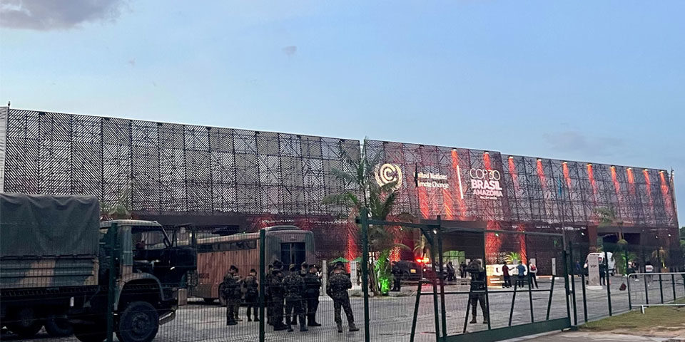 COP30_military_presence_960 Behind a green mesh security fence military personnel can be seen in combat fatigures and berets. A troop carrying lorry is parked to the right. In the background the COP30 convention centre with its three storey mesh wall and UNFCCC logo.