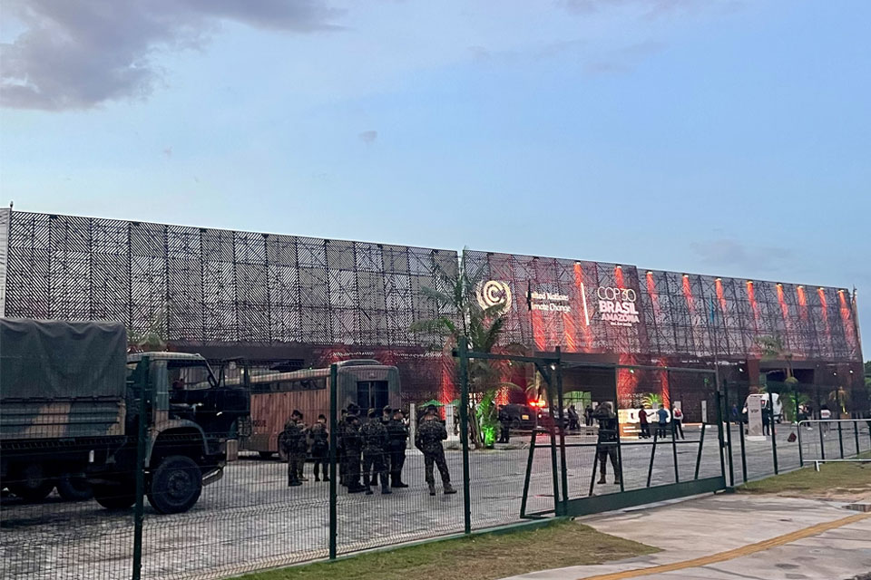 Behind a green mesh security fence military personnel can be seen in combat fatigures and berets. A troop carrying lorry is parked to the right. In the background the COP30 convention centre with its three storey mesh wall and UNFCCC logo.