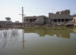 The top half of the image is a ruined building, the top floor has collapsed leaving piles of rubble, the bottom half of the image is brown floodwater, with the ruins reflected in it.