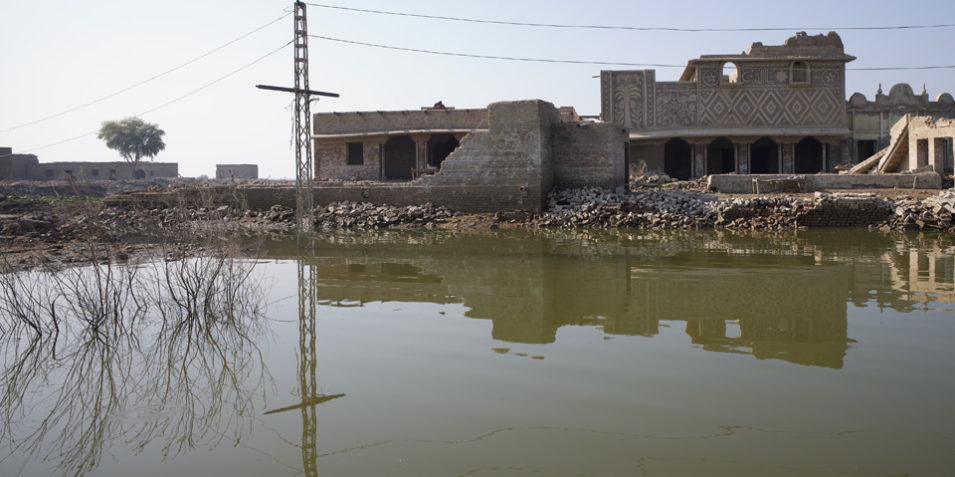 The top half of the image is a ruined building, the top floor has collapsed leaving piles of rubble, the bottom half of the image is brown floodwater, with the ruins reflected in it.