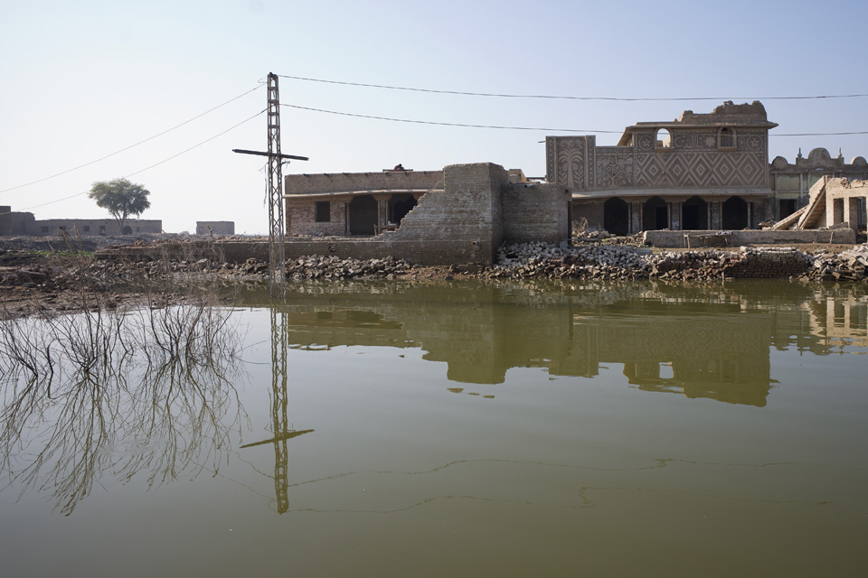The top half of the image is a ruined building, the top floor has collapsed leaving piles of rubble, the bottom half of the image is brown floodwater, with the ruins reflected in it.