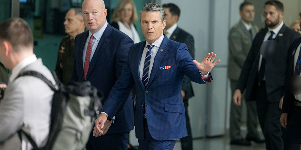 Hegseth_NATO_960 Pete Hegseth raises a hand palm out as he and male colleagues walk through a NATO meeting.