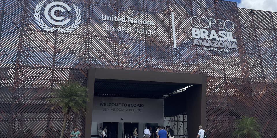The entrance to the COP3 exhibition centre in Belem, the large doorway is towered over by a metal mesh backdrop with the event and UNFCCC logos on them.