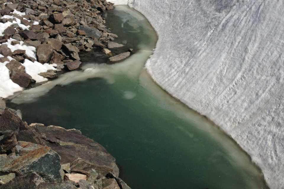 A close up view of a glacial lake shore. To the left rocky scree with a few patches of snow, in the centre, a patch of greenish water, it's fairly clear with ice around the edges. To the right, a striated slope of snow and ice descends into the water.