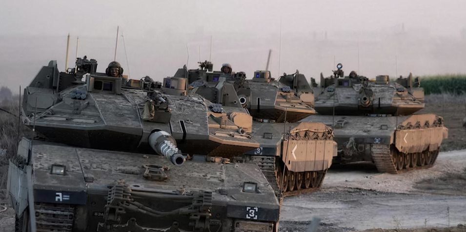 Four Israeli Merkava tanks drive along a sandy track, it is at dawn or dusk, the background is hazy with dust.