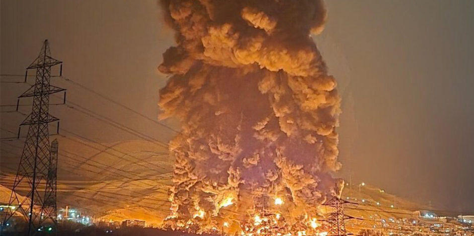 Nighttime image, a huge fire a smoke plume can be seen, glowing orange as a column of black smoke rises into the sky.