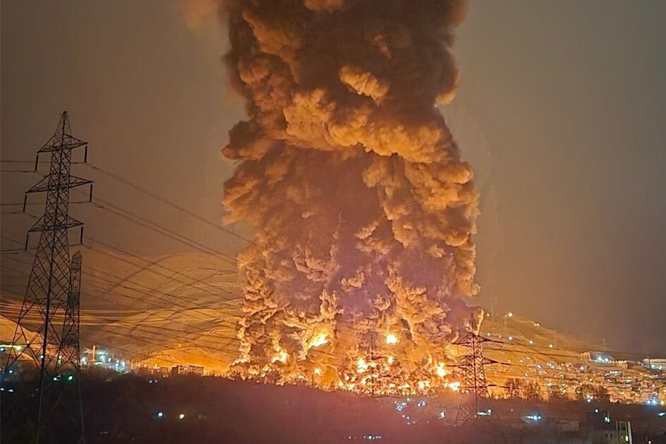 Nighttime image, a huge fire a smoke plume can be seen, glowing orange as a column of black smoke rises into the sky.