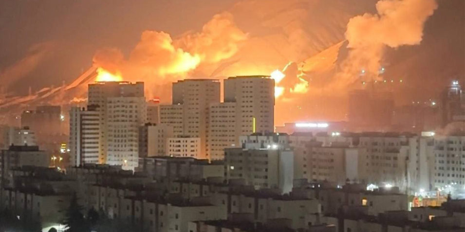 Night time. In the background the slopes of mountains, orange explosions and smoke plumes can be seen illuminating them, in the foreground many apartment blocks.