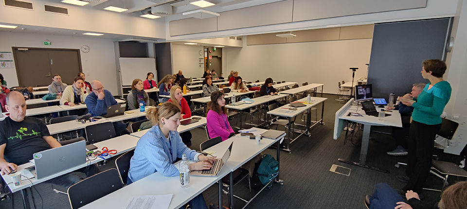 A classroom setting, around 20 people are sittign behind grey desks listening to a woman speak.