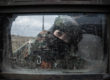 A soldier in camouflage and a helmet peers into a mud speckled vehicle window, their face is covered and only their eyes can be seen.
