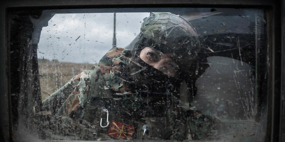 A soldier in camouflage and a helmet peers into a mud speckled vehicle window, their face is covered and only their eyes can be seen.