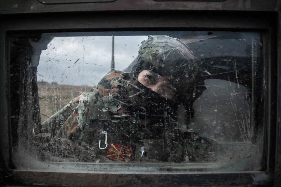 A soldier in camouflage and a helmet peers into a mud speckled vehicle window, their face is covered and only their eyes can be seen.