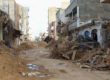 View up a heavily damaged street. Building frontages have collapsed, piles of sediment and debris have been pushed to the side to create a path.