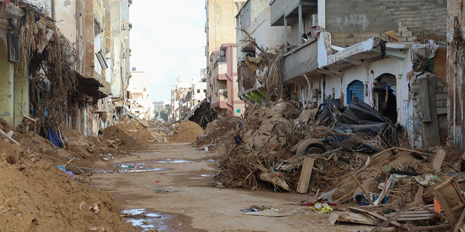 View up a heavily damaged street. Building frontages have collapsed, piles of sediment and debris have been pushed to the side to create a path.